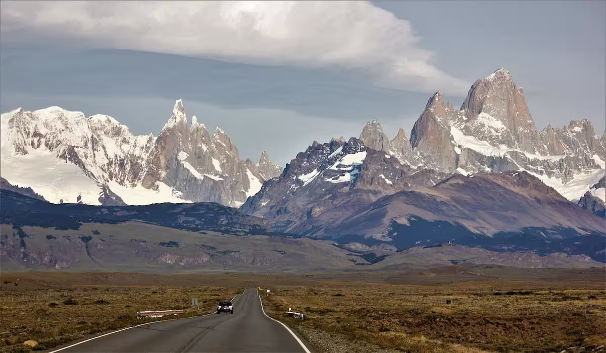 Ruta de asfalto recta que cruza una llanura de pastizal seco, con un cordón de picos graníticos nevados en el horizonte
